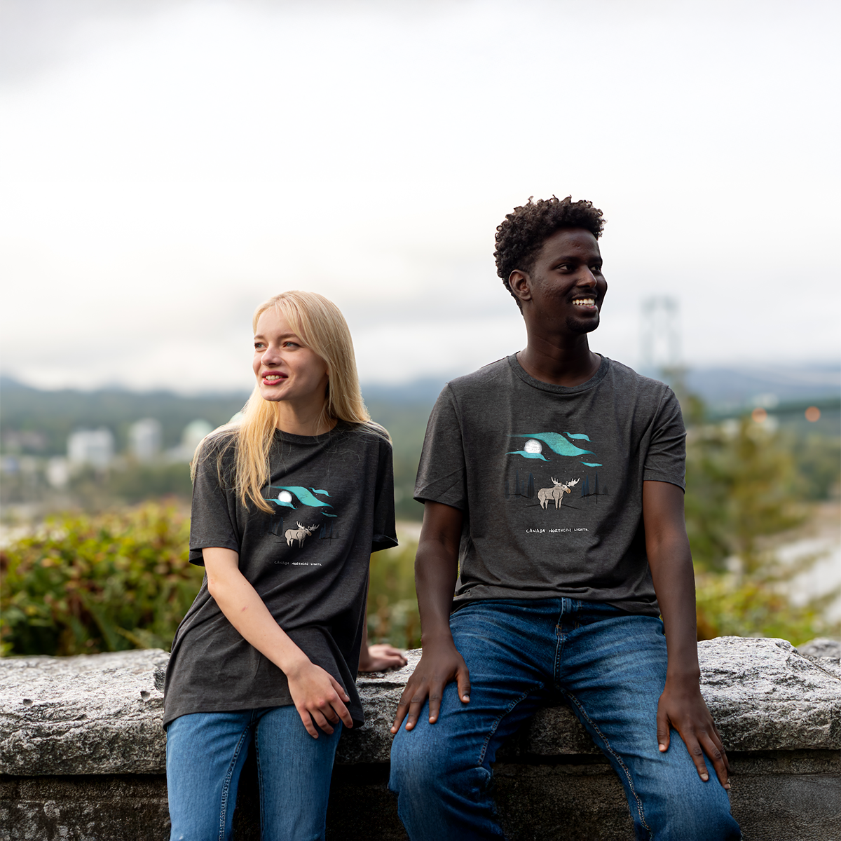 Two models smiling and sitting by a stone ledge wearing dark grey VELU T-shirts featuring a moose under the northern lights. Made sustainably in Vancouver, Canada.