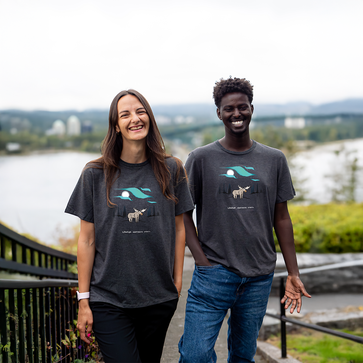 Smiling models wearing dark grey “Aurora Horizon” VELU T-shirts with northern lights and moose design, captured outdoors in Vancouver. Ethically made in Canada.