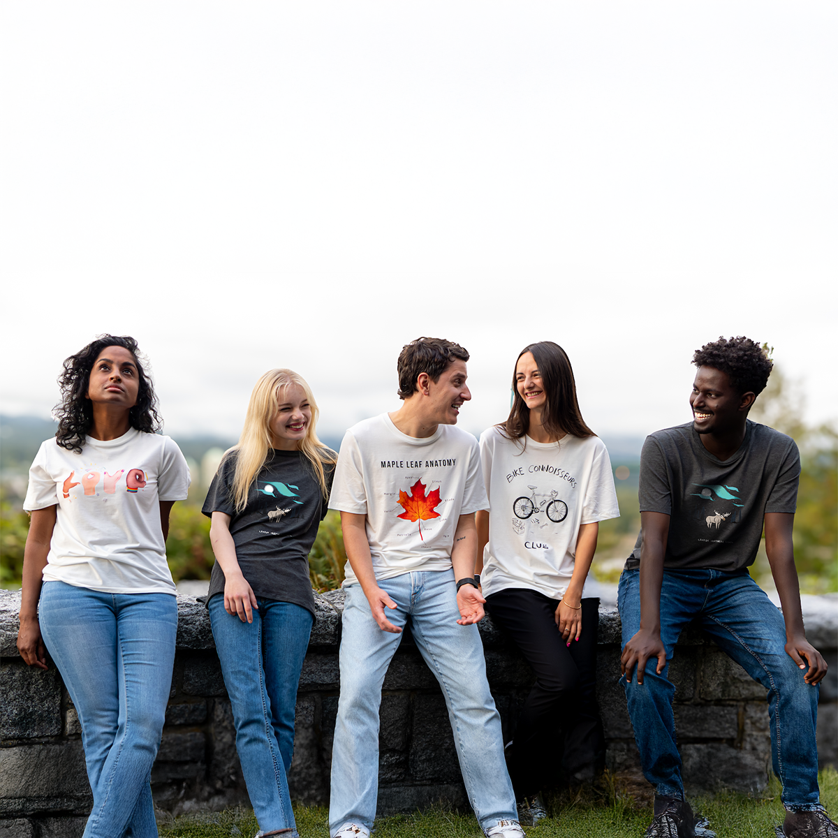 Five diverse models laughing and wearing different VELU t-shirts — including Love in Hands, Maple Leaf Anatomy, Bike Connoisseurs Club, and Aurora Horizon — shot outdoors in Canada. Ethical, sustainable fashion made in Vancouver.