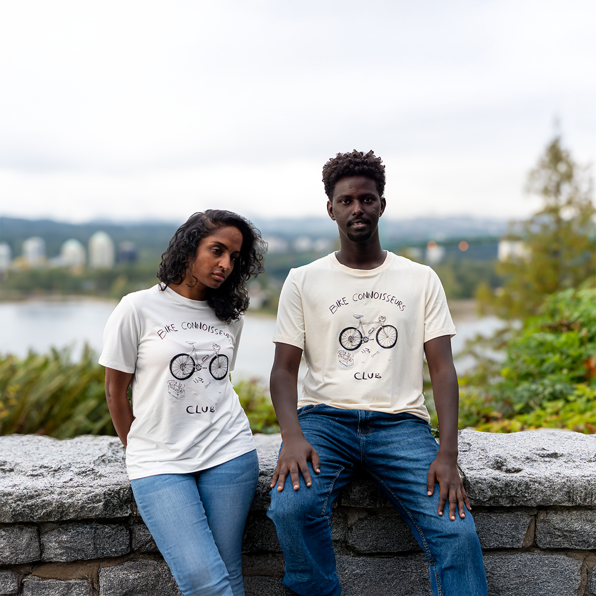 Two people wearing “Bike Connoisseurs Club” graphic T-shirts by VELU, sitting on a stone wall with a city skyline and lake in the background. Ethically made in Canada using sustainable lyocell cotton blend.