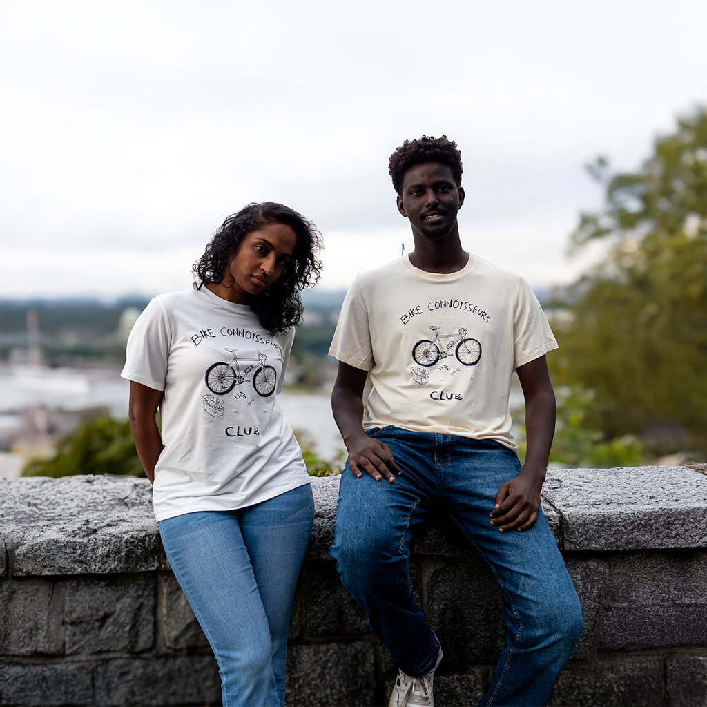 Two people wearing VELU t-shirts with hand-drawn bicycle illustrations and the words “Bike Connoisseurs Club”, standing on a stone wall with a scenic Canadian landscape in the background. Made in Vancouver.