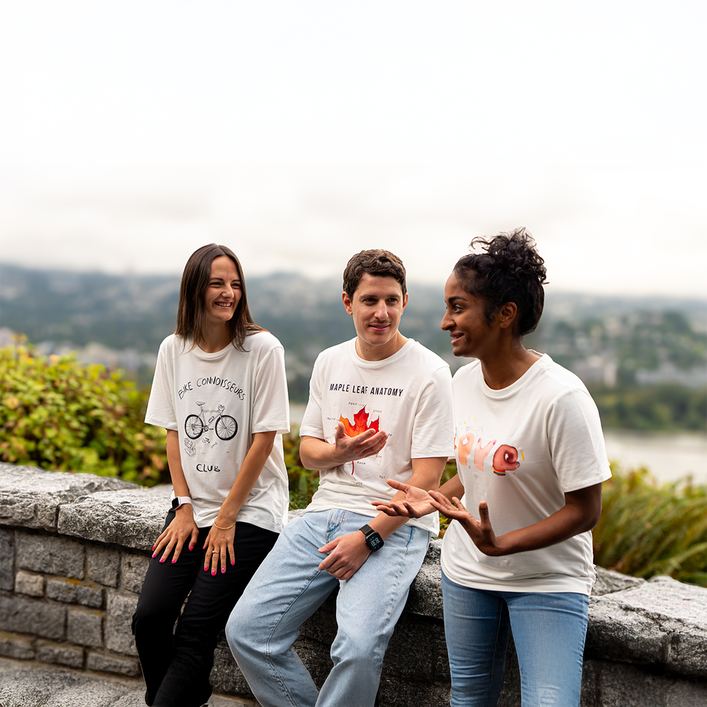 Three people wearing VELU’s sustainable t-shirts — featuring the “Bike Connoisseurs Club,” “Maple Leaf Anatomy,” and “Love in Hands” designs — casually chatting outdoors with the Canadian landscape in the background. Made in Vancouver.