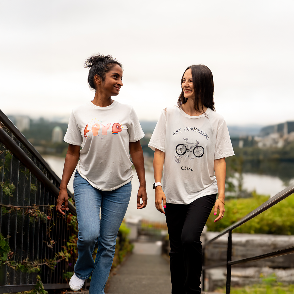 Two women wearing VELU’s “Love in Hands” and “Bike Connoisseurs Club” t-shirts walking outdoors on a scenic path overlooking the Canadian landscape. Both styles are made in Vancouver using sustainable materials.
