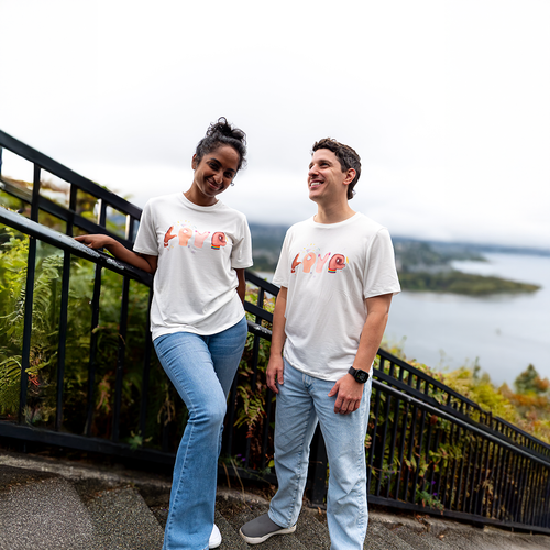 Two people wearing white VELU t-shirts with “LOVE” printed in hand-shaped letters, standing on a scenic path overlooking nature and water. Made ethically in Vancouver, Canada.