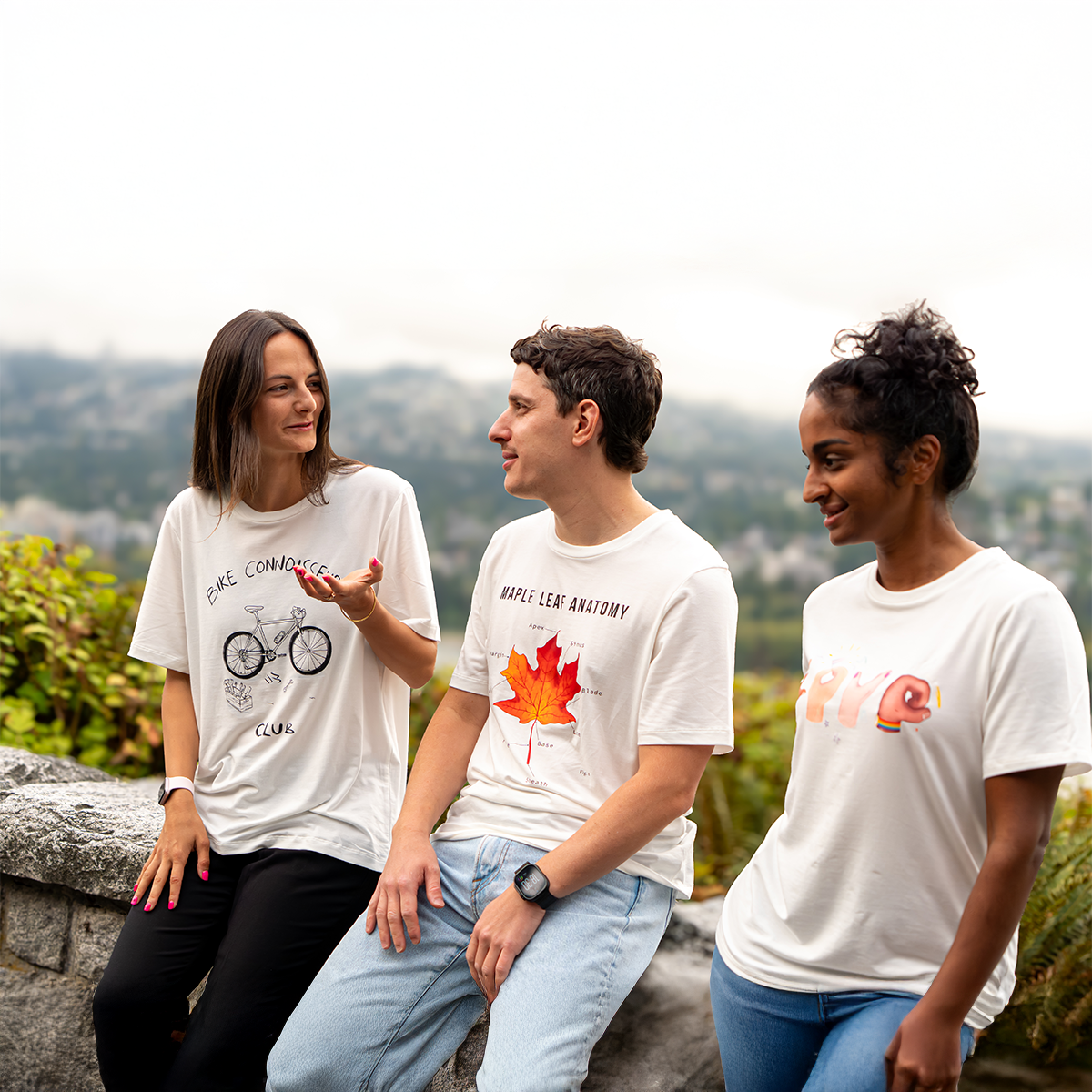 Three friends wearing VELU T-shirts (“Bike Connoisseurs,” “Maple Leaf Anatomy,” and “Love in Hands”) laughing and chatting outdoors in Vancouver. Canadian-made, sustainable apparel.