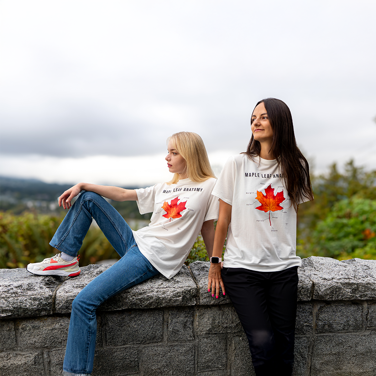 Two women wearing “Maple Leaf Anatomy” graphic T-shirts by VELU, seated on a stone wall with a scenic view of mountains and trees in the background. Made in Canada with bamboo rayon blend.