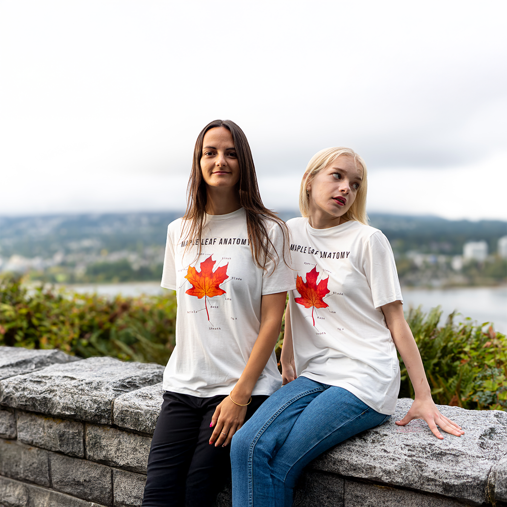 Two women wearing “Maple Leaf Anatomy” T-shirts by VELU, sitting on a stone wall with scenic forest and lake views in the background.