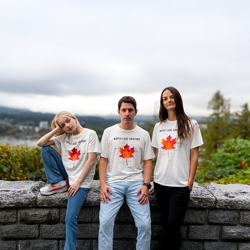 Three people wearing white “Maple Leaf Anatomy” T-shirts standing outdoors in front of a scenic Canadian landscape with mountains and trees.