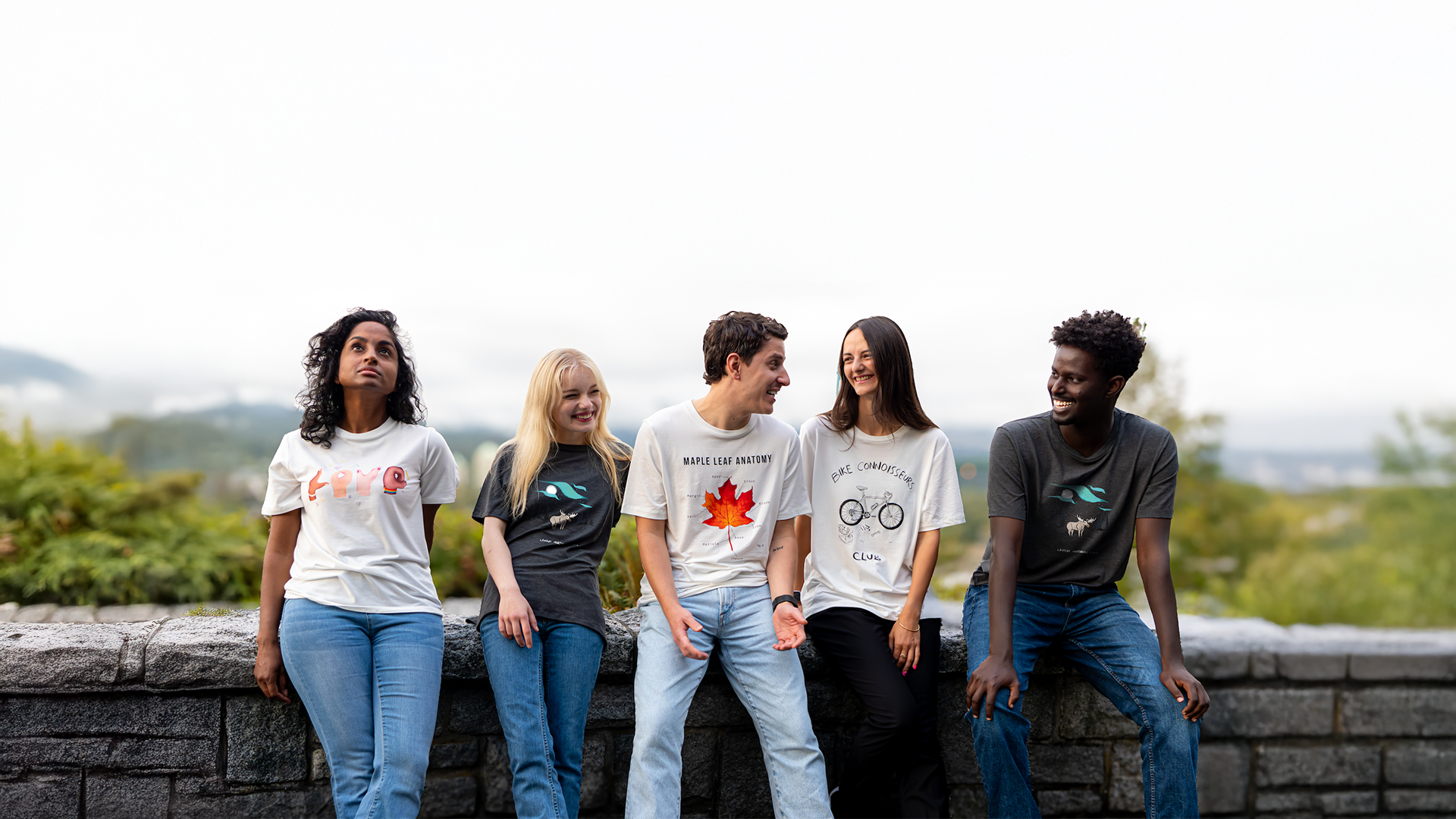 Five people wearing sustainably made Canadian t-shirts, smiling and sitting on a stone wall with a scenic forest and lake background.